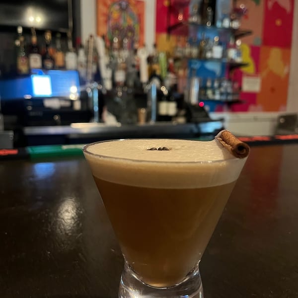 A martini glass with a frothy, light brown drink, garnished with coffee beans and a cinnamon stick.