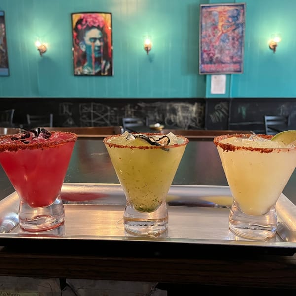 Three colorful margaritas in stemmed glasses on a silver tray, with a teal wall and artwork in the background.