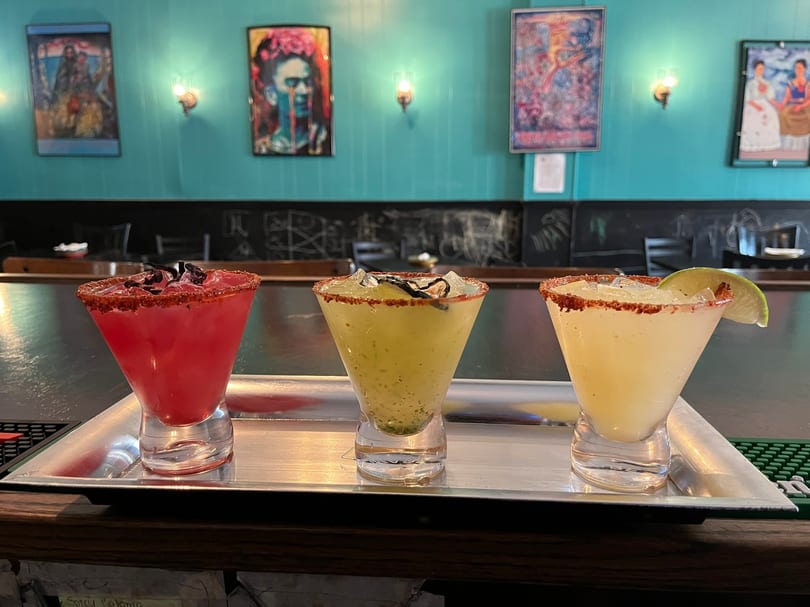 Three colorful margaritas in stemmed glasses on a silver tray, with a teal wall and artwork in the background.