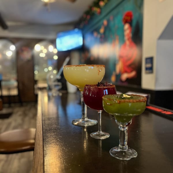 Three colorful margaritas, yellow, red, and green, sit on a dark bar with a vibrant mural in the background.