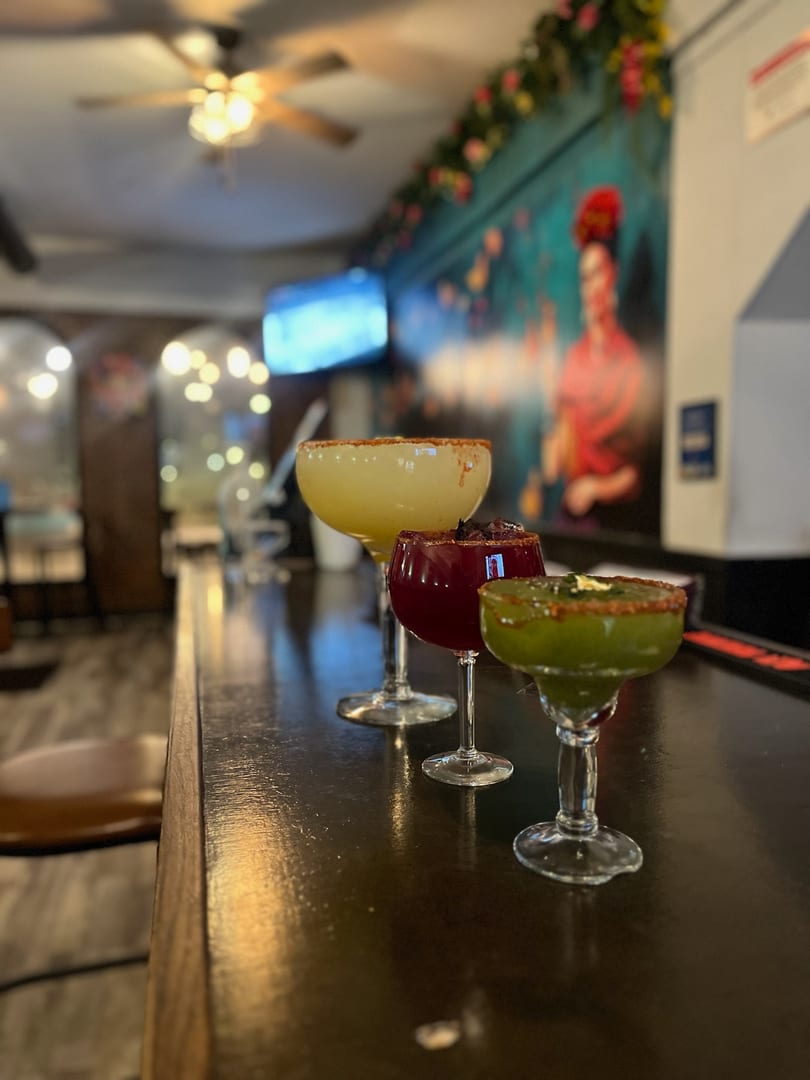 Three colorful margaritas, yellow, red, and green, sit on a dark bar with a vibrant mural in the background.