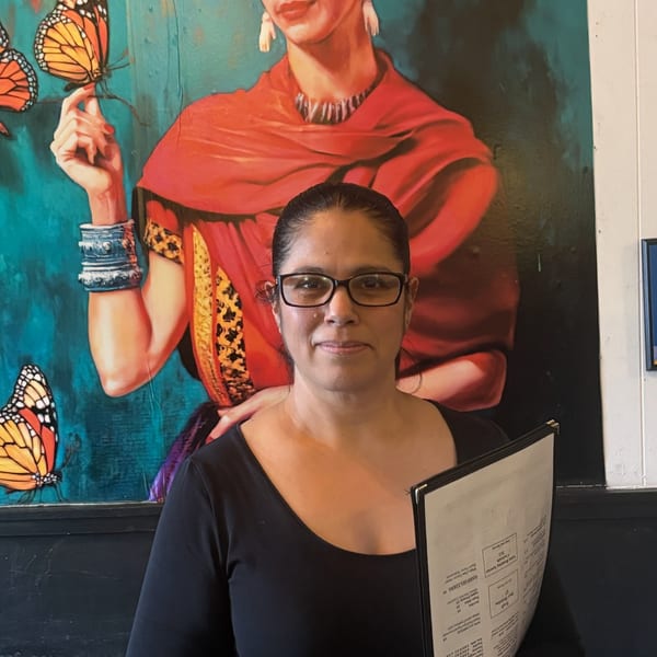 Woman in glasses holding a menu stands before a vibrant Frida Kahlo mural with butterflies.