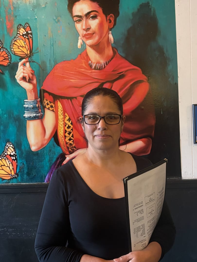 Woman in glasses holding a menu stands before a vibrant Frida Kahlo mural with butterflies.