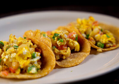 Three breakfast tacos filled with scrambled eggs, vegetables, and possibly cheese, served on a white plate. The tacos are made with golden corn tortillas and are arranged in a row.