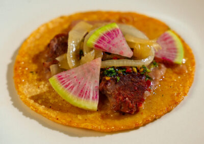 A corn tortilla topped with grilled meat, sautéed onions, and slices of watermelon radish, served on a white plate.