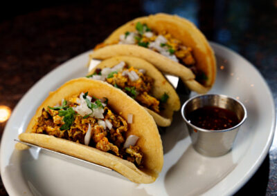 Three soft shell tacos filled with seasoned meat, scrambled eggs, chopped onions, and cilantro are served on a white plate with a small metal cup of red salsa.