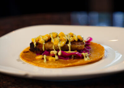 A taco on a white plate topped with grilled tofu, corn, shredded red cabbage, and drizzled with a creamy yellow sauce, all on a yellow corn tortilla.