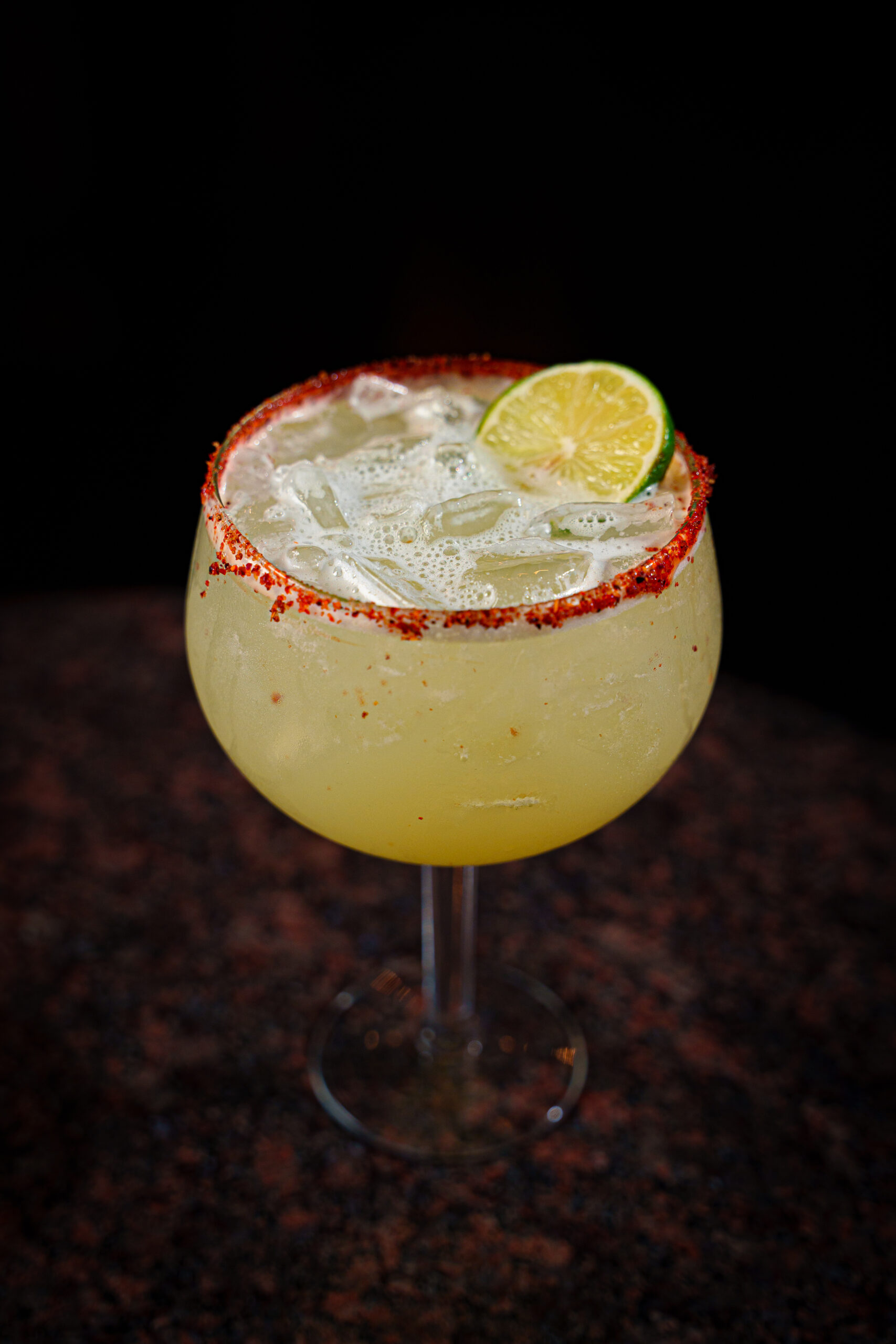 A large margarita glass filled with a yellow cocktail, topped with ice, garnished with a lime slice, and a chili powder rim, set against a dark background.