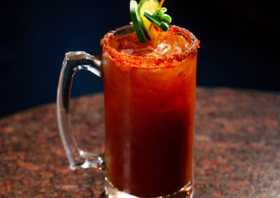 A glass mug filled with a reddish-brown drink, garnished with lime slices, a jalapeño slice, and a chili powder rim, sits on a dark speckled surface against a dark background.