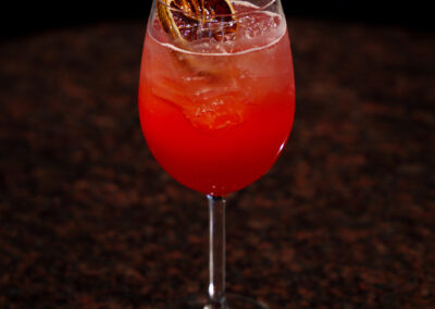 A vibrant red cocktail in a wine glass filled with ice, garnished with a dried citrus wheel, sits on a dark, speckled stone surface.