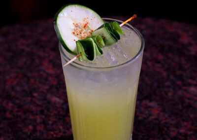 A tall glass of yellow-green cocktail with ice, garnished with a cucumber slice and folded cucumber ribbon on a skewer, sits on a dark marbled surface against a black background.