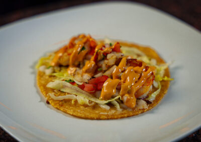 A crispy taco topped with lettuce, diced tomatoes, grilled shrimp, and drizzled with orange sauce, served on a white plate.