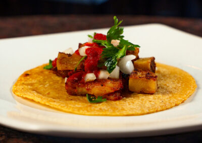 A taco on a yellow corn tortilla topped with roasted potatoes, onions, red salsa, and fresh cilantro, served on a white plate.